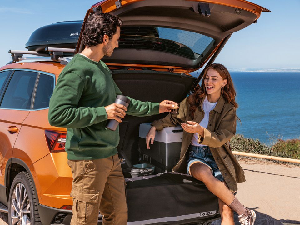 Ein Mann und eine Frau stehen hinter einem orangen Škoda mit geöffneter Heckklappe. Die Frau sitzt auf der Kofferraumkante und schaut auf ihr Handy, während der Mann eine Thermoskanne hält und mit ihr spricht. Im Hintergrund sieht man das Meer.