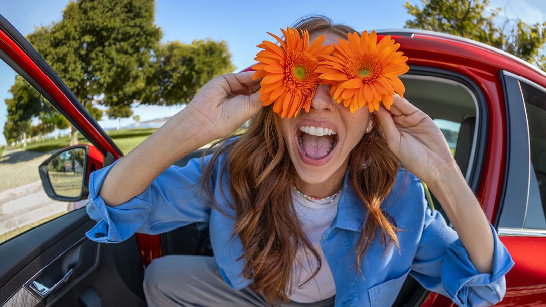 Eine Frau sitzt lachend in einem roten Škoda und hält sich zwei orangefarbene Blumen an die Augen.