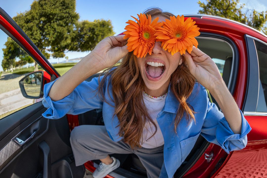 Eine Frau sitzt lachend in einem roten Škoda und hält sich zwei orangefarbene Blumen an die Augen.