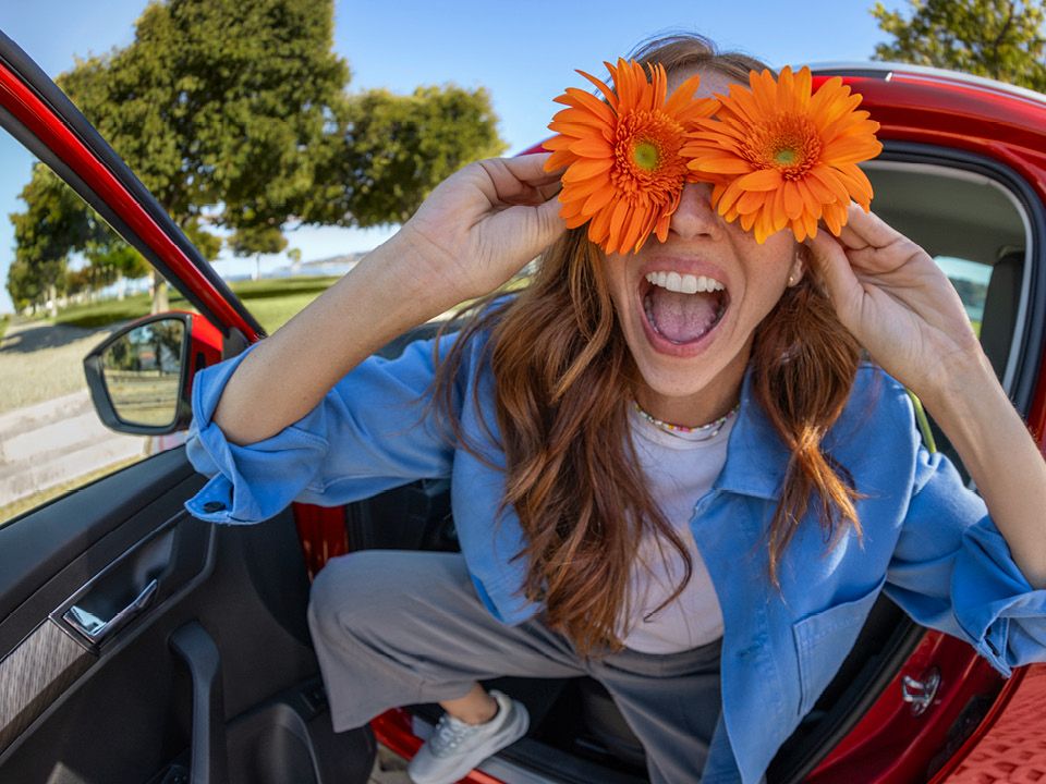 Eine Frau sitzt lachend in einem roten Škoda und hält sich zwei orangefarbene Blumen an die Augen.
