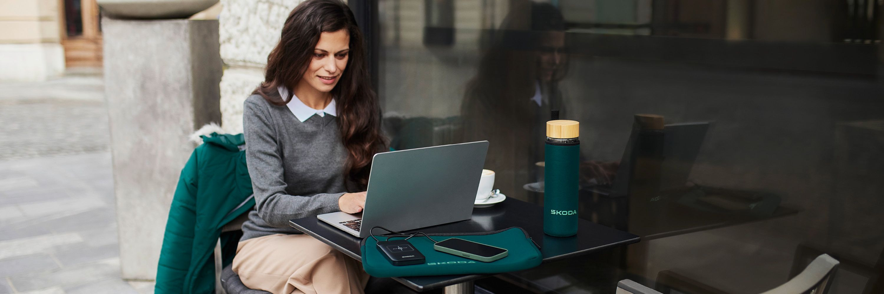 Eine Frau mit Laptop sitzt vor einem Cafe. Auf dem Tisch liegt eine grüne Laptop Hülle mit Škoda Schriftzug. Auf dem Tisch eine grüne Škoda Glasflasche.