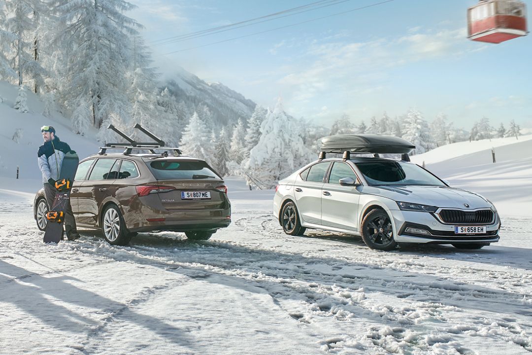 Ein brauner Škoda Octavia mit Dachträger und ein silberner Škoda Superb mit Dachbox parken in einer winterlichen Landschaft. Ein Mann mit Snowboard steht neben den Fahrzeugen.