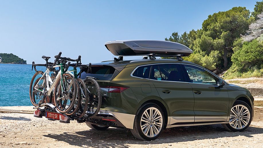 Ein Škoda Enyaq in Olibo-Grün Metallic mit Fahrradheckträger und Dachbox parkt in einer Bucht am Meer.
