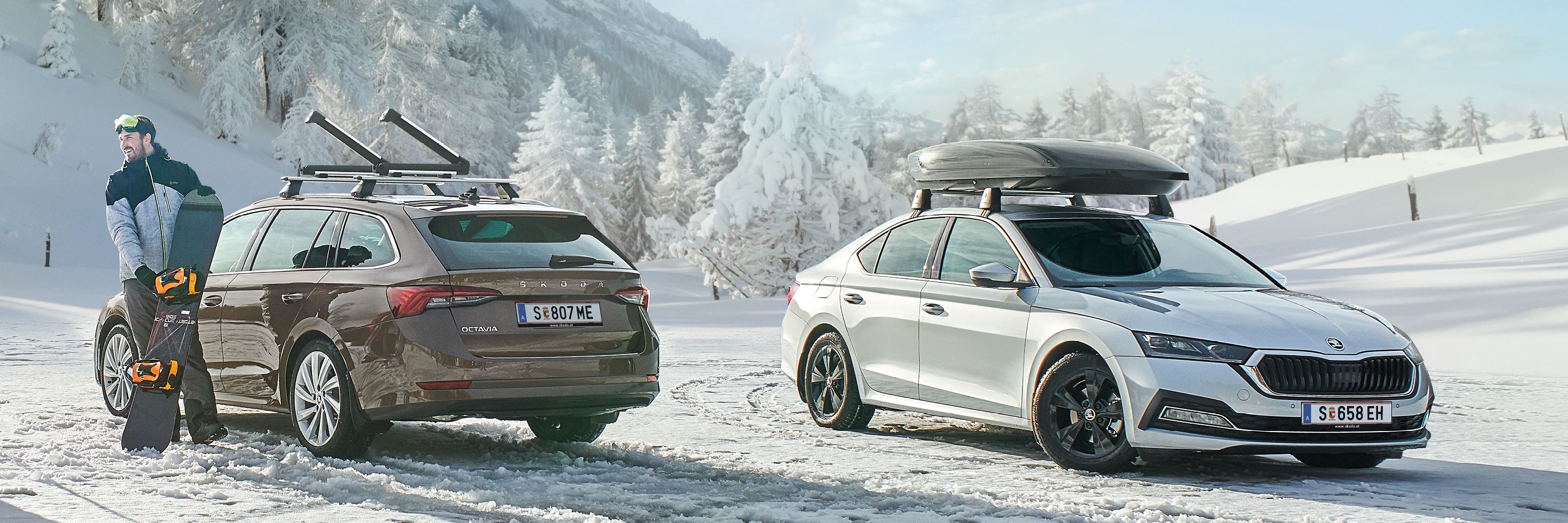 Ein brauner Škoda Octavia mit Dachträger und ein silberner Škoda Superb mit Dachbox parken in einer winterlichen Landschaft. Ein Mann mit Snowboard steht neben den Fahrzeugen.