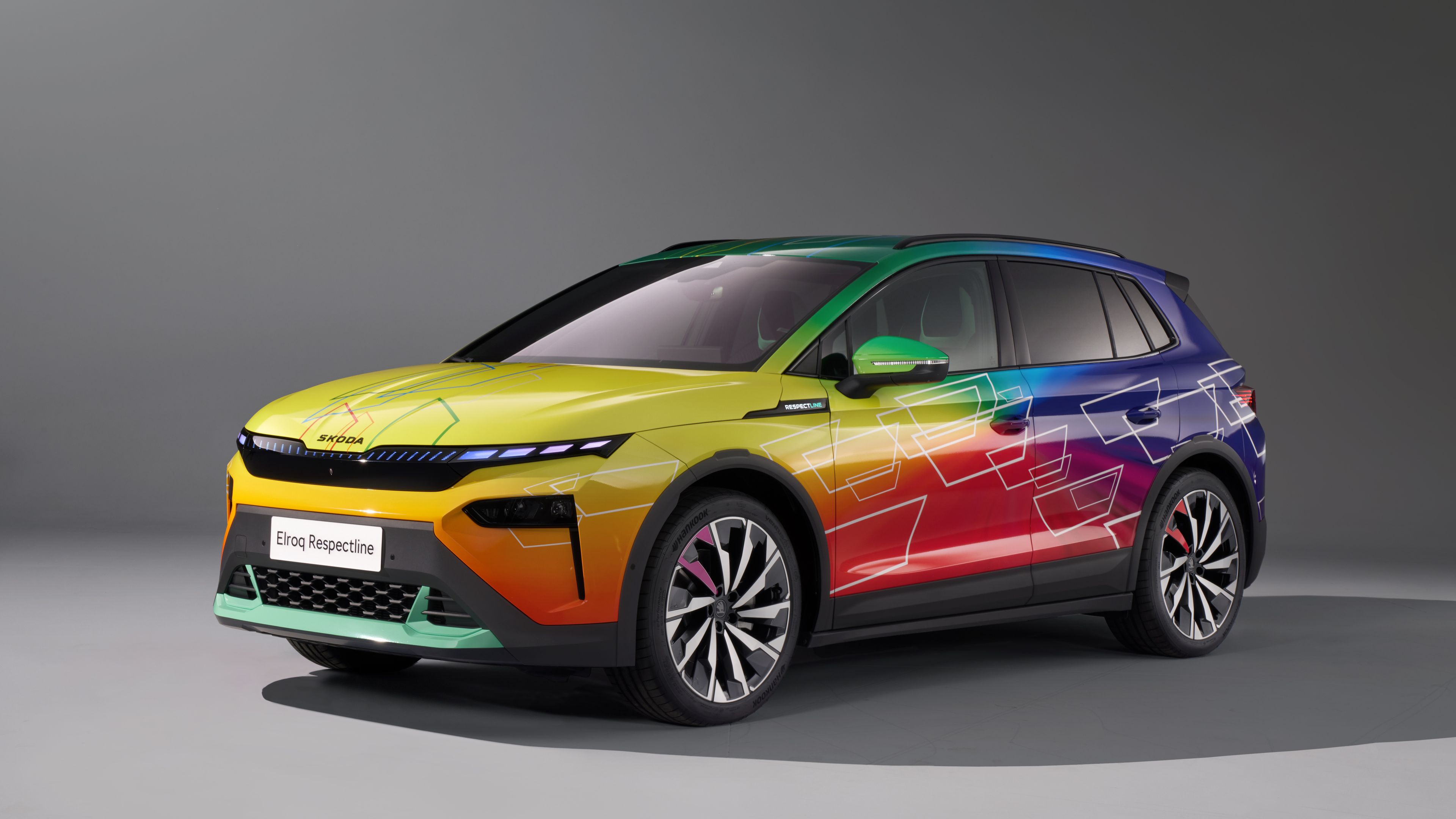 Ein Škoda Elroq im Studio, lackiert in Regenbogen-Farbverlauf von Gelb über Grün bis Violett. Die Karosserie ist zusätzlich mit weißen geometrischen Linien und Kästen verziert. Auf dem Nummernschild steht „Elroq Respectline“.