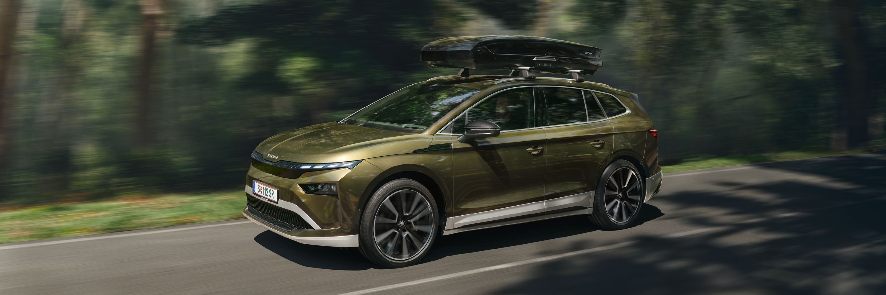 Ein Škoda  Enyaq mit Transportbox auf dem Dach fährt auf einer Straße durch einen Wald.