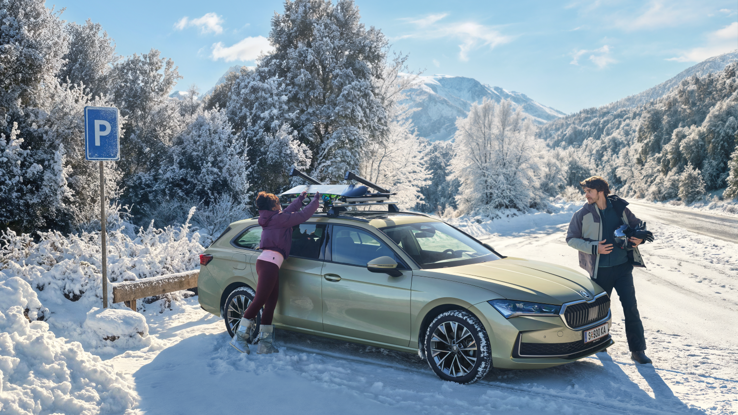 Ein hellgrüner Škoda Suberb Kombi steht auf einem verschneiten Parkplatz in einer winterlichen Berglandschaft, umgeben von schneebedeckten Bäumen. Eine Person lädt Skiausrüstung auf den Dachträger, während eine andere daneben steht und etwas in den Händen hält.