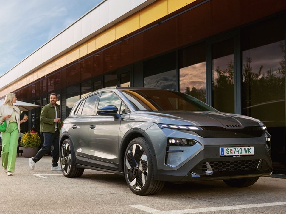 Ein Mann und eine Frau verabschieden sich hinter einem geparkten, grauen Škoda Elroq.