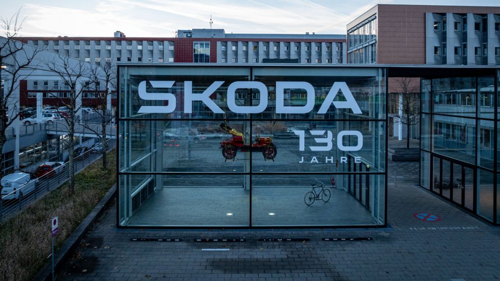 Das Bild zeigt eine moderne Glaskonstruktion mit dem Schriftzug "SKODA 130 JAHRE" darauf. Im Inneren der Konstruktion befinden sich ein rotes Motorrad und ein Fahrrad. Im Hintergrund sieht man Gebäude und einen bewölkten Himmel.