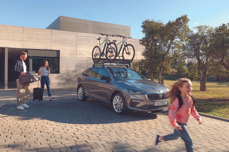 Ein grauer Škoda Scala mit Fahrraddachträger parkt vor einem Haus, die Eltern bereiten sich mit ihrer Tochter auf die Fahrt vor.