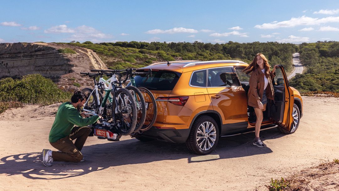 Ein orangefarbener Škoda Kodiaq steht auf einem sandigen Weg. Zwei Fahrräder sind auf einem Fahrradträger am Heck des Autos befestigt. Eine Person in grüner Kleidung überprüft den Fahrradträger, während eine Person in brauner Kleidung aus dem Fahrzeug steigt