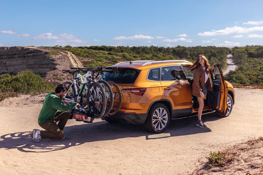 Ein orangefarbener Škoda Kodiaq steht auf einem sandigen Weg. Zwei Fahrräder sind auf einem Fahrradträger am Heck des Autos befestigt. Eine Person in grüner Kleidung überprüft den Fahrradträger, während eine Person in brauner Kleidung aus dem Fahrzeug steigt
