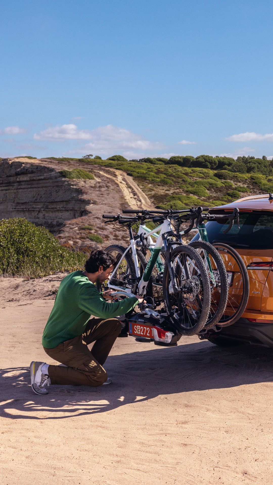 Ein orangefarbener Škoda Kodiaq steht auf einem sandigen Weg. Zwei Fahrräder sind auf einem Fahrradträger am Heck des Autos befestigt. Eine Person in grüner Kleidung überprüft den Fahrradträger, während eine Person in brauner Kleidung aus dem Fahrzeug steigt