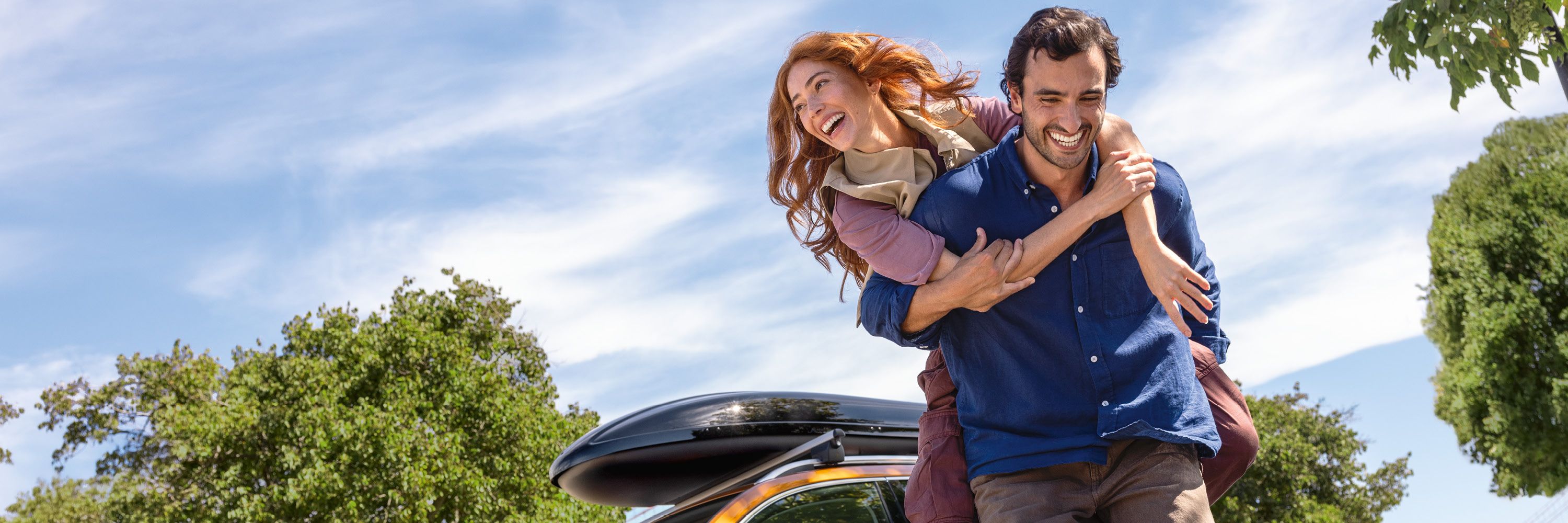 Ein junger Mann trägt seine Partnerin auf dem Rücken. Im Hintergrund ist ein oranger Škoda Karoq mit Dachbox auf dem Dach zu sehen.