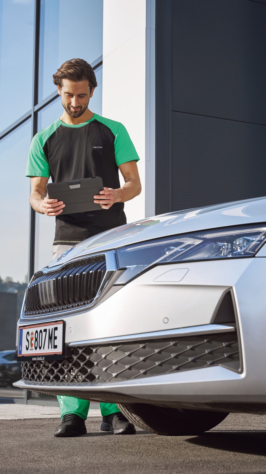 Ein Škoda Mechaniker steht neben einem Škoda und blickt auf ein Tablet.