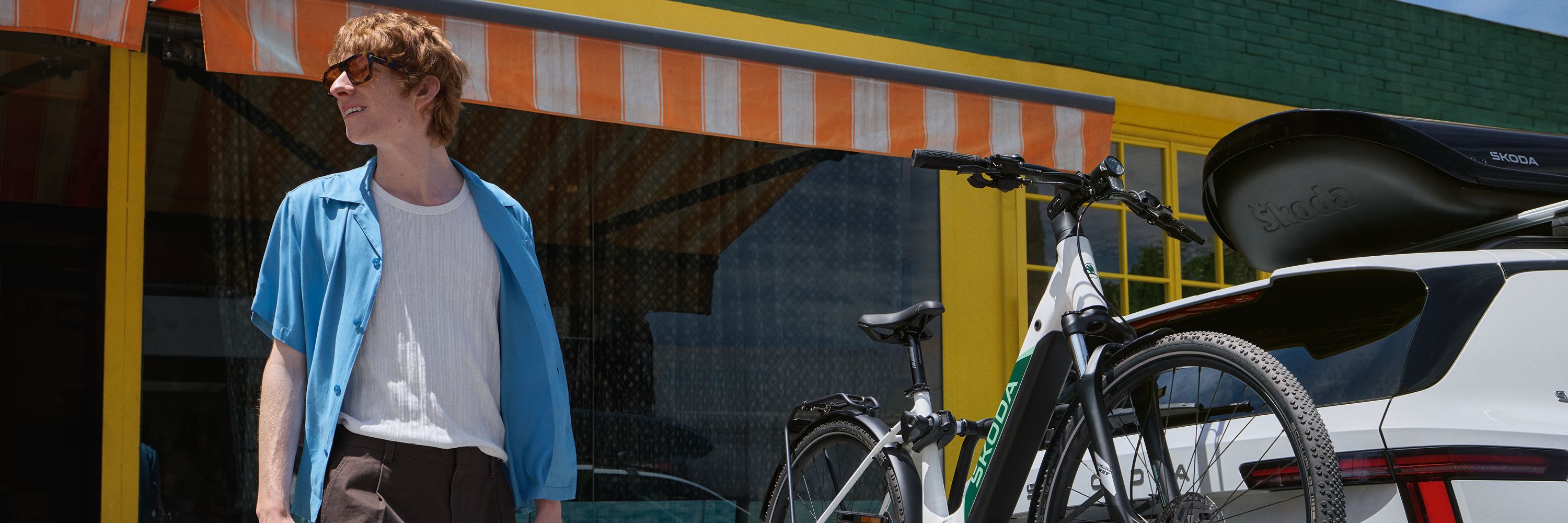 Junger Mann mit lockigen roten Haaren steht lächelnd neben einem weißen Škoda Epiq, an dessen Heck ein E-Bike auf einem Fahrradträger montiert ist.