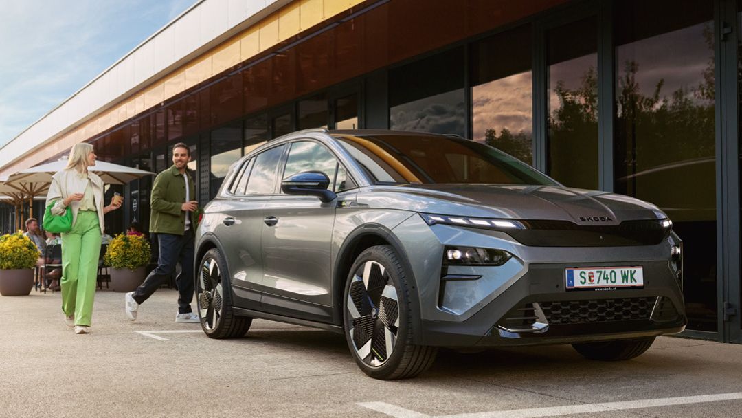 Ein Mann und eine Frau verabschieden sich hinter einem geparkten, grauen Škoda Elroq.