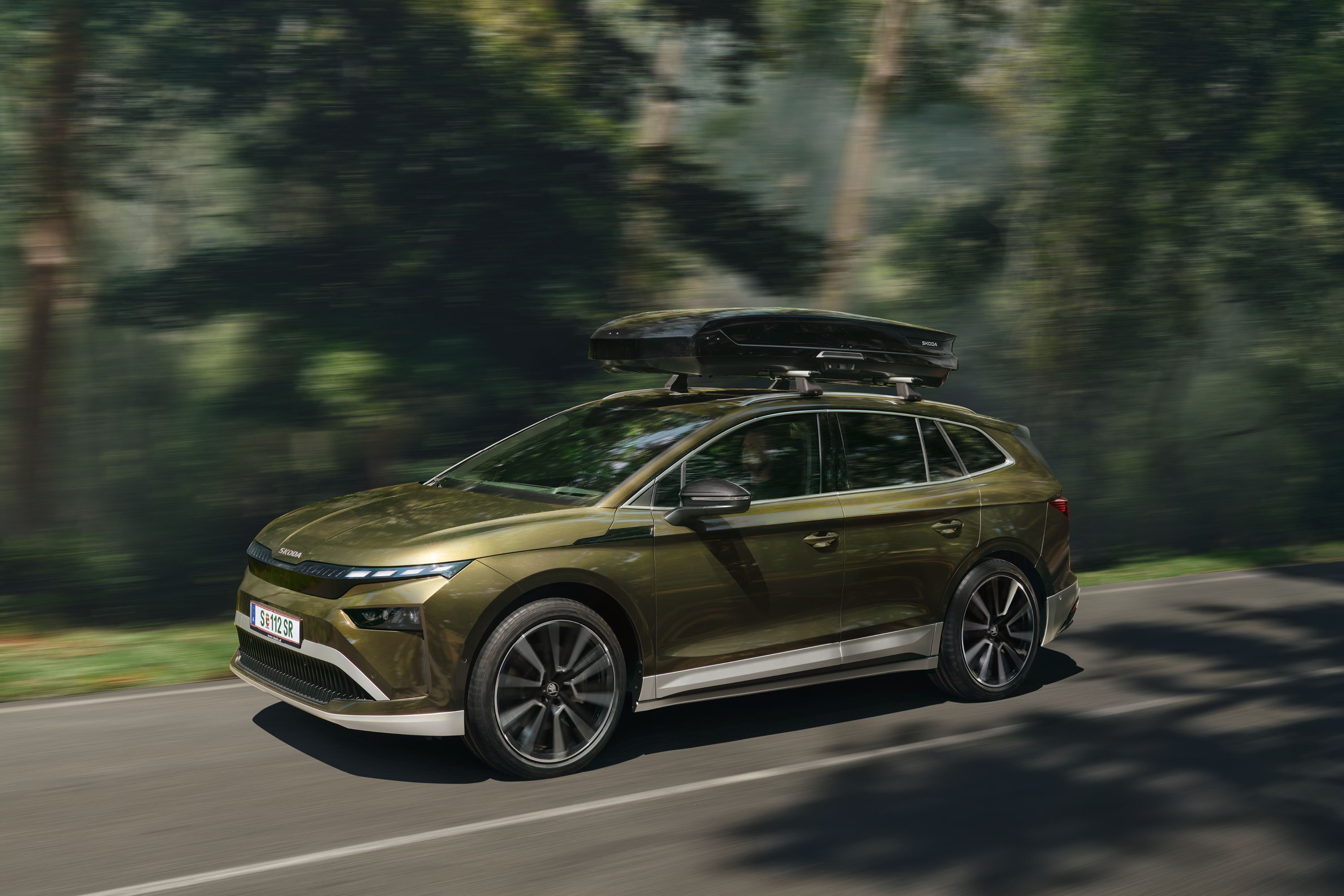 Ein Škoda  Enyaq mit Transportbox auf dem Dach fährt auf einer Straße durch einen Wald.