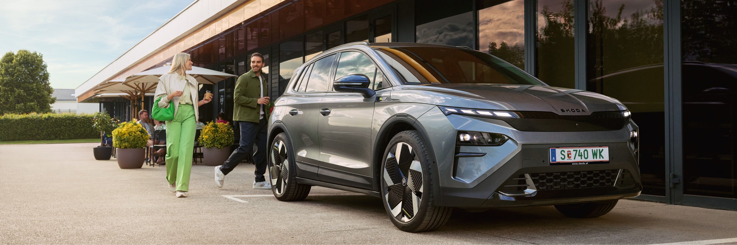 Ein Mann und eine Frau verabschieden sich hinter einem geparkten, grauen Škoda Elroq.