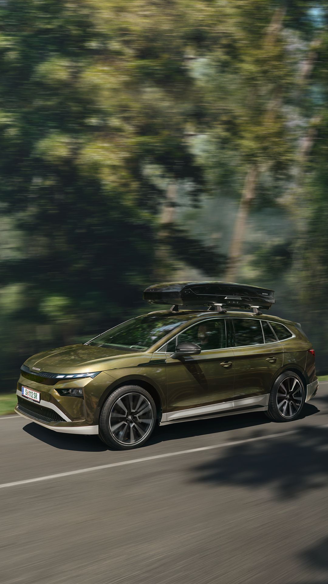 Ein Škoda  Enyaq mit Transportbox auf dem Dach fährt auf einer Straße durch einen Wald.