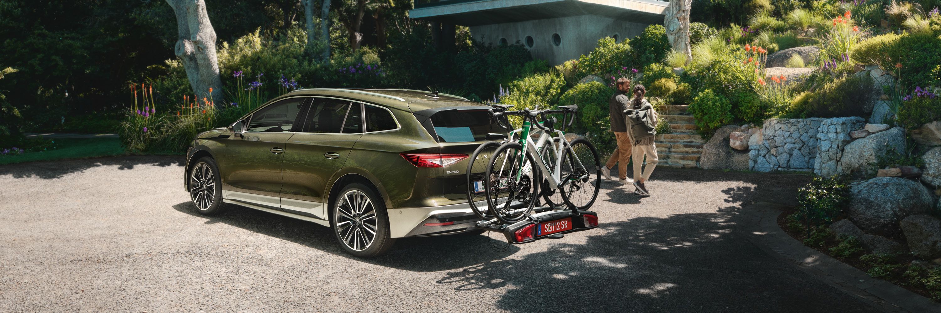 Ein grüner Škoda Enyaq mit zwei Fahrrädern auf einem Fahrradträger ist vor einem modernen Haus geparkt. Zwei Personen laufen darauf zu. Die Umgebung ist von Bäumen und Pflanzen umgeben.