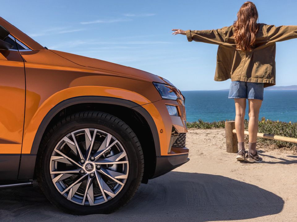 Eine Person mit langen Haaren steht mit ausgebreiteten Armen am Rand einer Klippe und blickt auf das Meer. Links im Vordergrund ist die Vorderseite eines orangefarbenen Škoda zu sehen. 