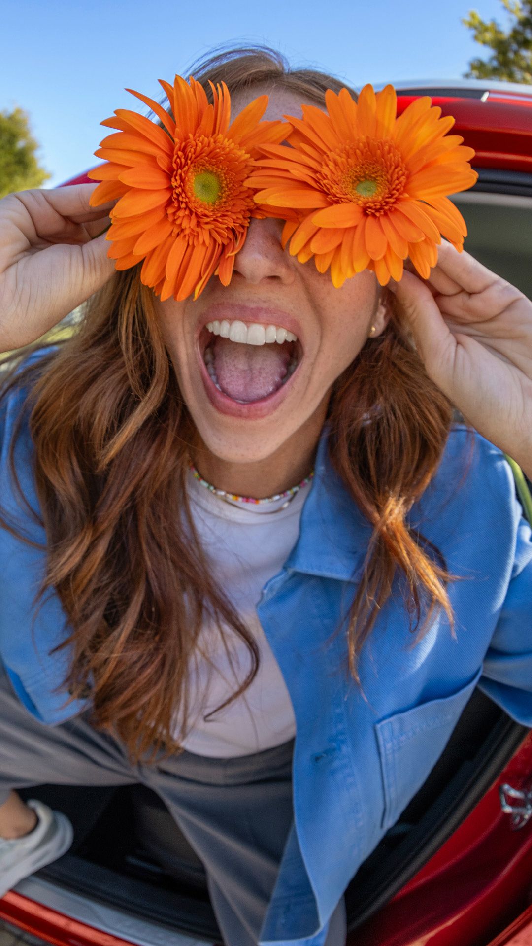 Eine Frau sitzt lachend in einem roten Škoda und hält sich zwei orangefarbene Blumen an die Augen.