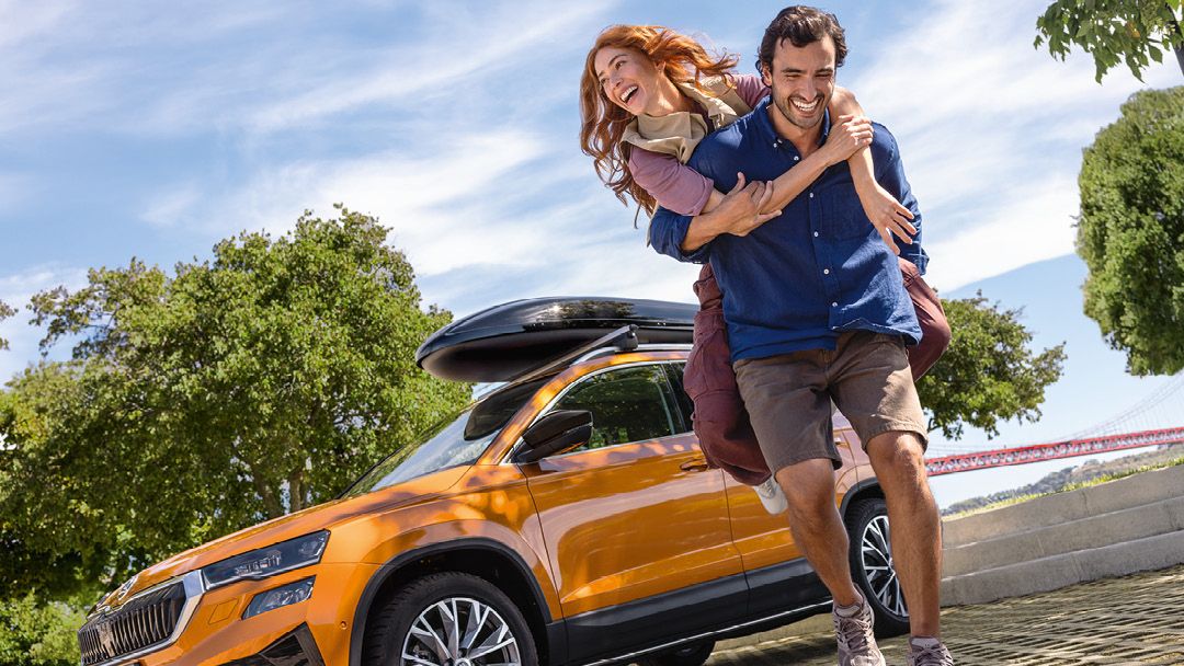Ein junger Mann trägt seine Partnerin auf dem Rücken. Im Hintergrund ist ein oranger Škoda Karoq mit Dachbox auf dem Dach zu sehen.