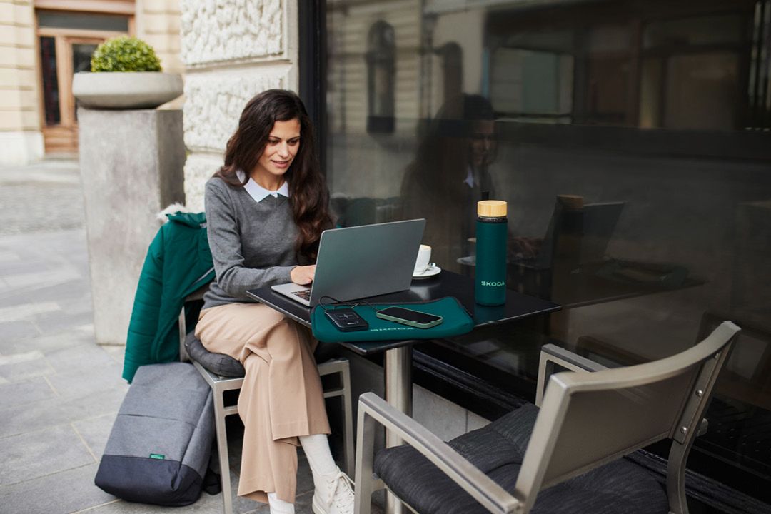 Eine Frau mit Laptop sitzt vor einem Cafe. Auf dem Tisch liegt eine grüne Laptop Hülle mit Škoda Schriftzug. Auf dem Tisch eine grüne Škoda Glasflasche.