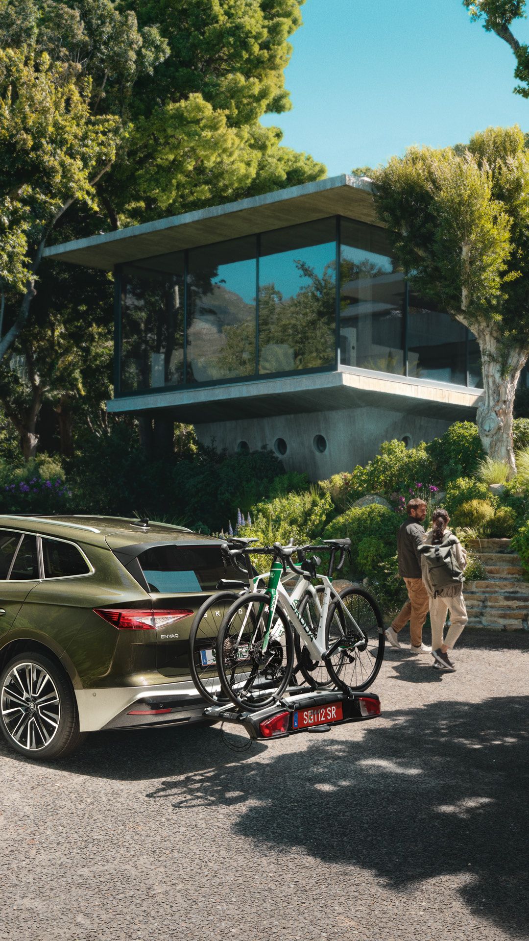 Ein grüner Škoda Enyaq mit zwei Fahrrädern auf einem Fahrradträger ist vor einem modernen Haus geparkt. Zwei Personen laufen darauf zu. Die Umgebung ist von Bäumen und Pflanzen umgeben.