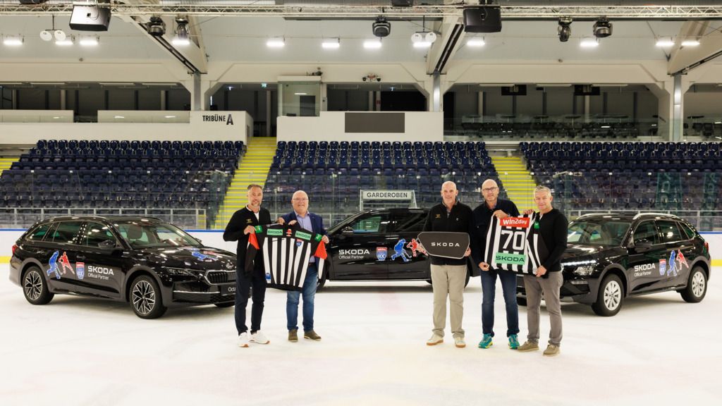 Fünf Personen stehen auf einer Eisbahn und halten verschiedene Gegenstände, darunter ein Schiedsrichtertrikot und ein Eishockeytrikot. Hinter ihnen sind drei schwarze Autos mit Sponsorenlogos geparkt. Die Tribünen im Hintergrund sind leer.