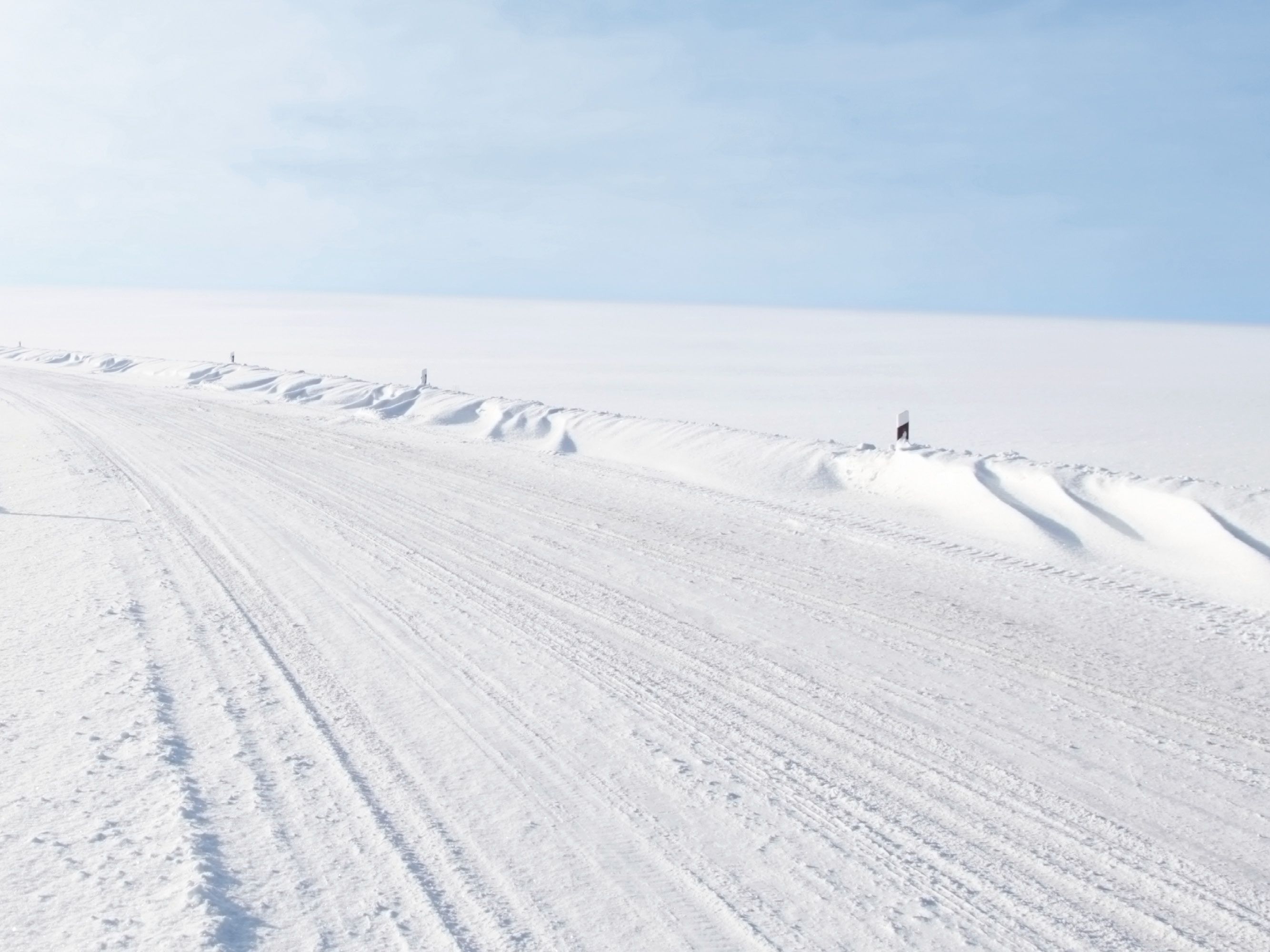 Weite, schneebedeckte Landschaft mit einer breiten, verschneiten Straße, die nach links aus dem Bild führt. Am Straßenrand türmen sich niedrige Schneeverwehungen.