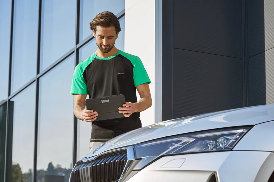 Ein Škoda Mechaniker steht neben einem Škoda und blickt auf ein Tablet.