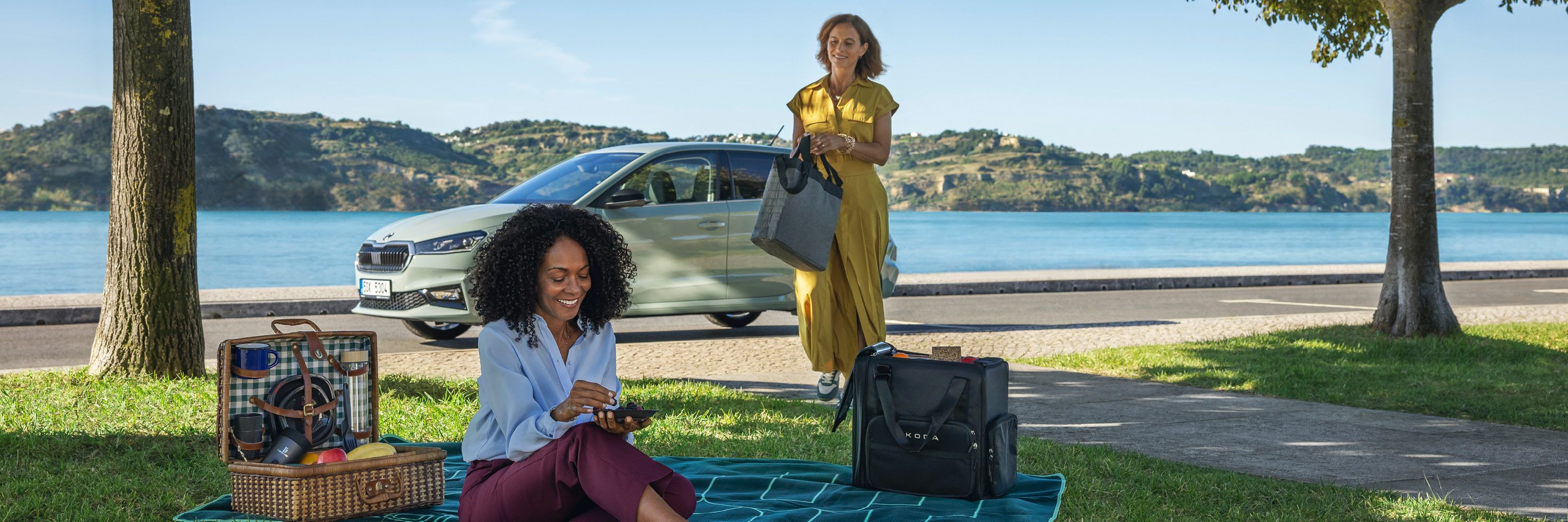 Zwei Frauen sind bei einem Picknick am See. Eine Frau sitzt auf einer Decke und isst, während die andere Frau im gelben Kleid einige Taschen trägt und sich der Decke nähert. Im Hintergrund ist ein geparkter Škoda und eine schöne Seelandschaft sichtbar.