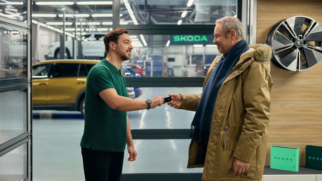 Ein Škoda Mitarbeiter übergibt die Autoschlüssel an einen Kunden. Im Hintergrund ist eine Škoda Werkstatt zu sehen.