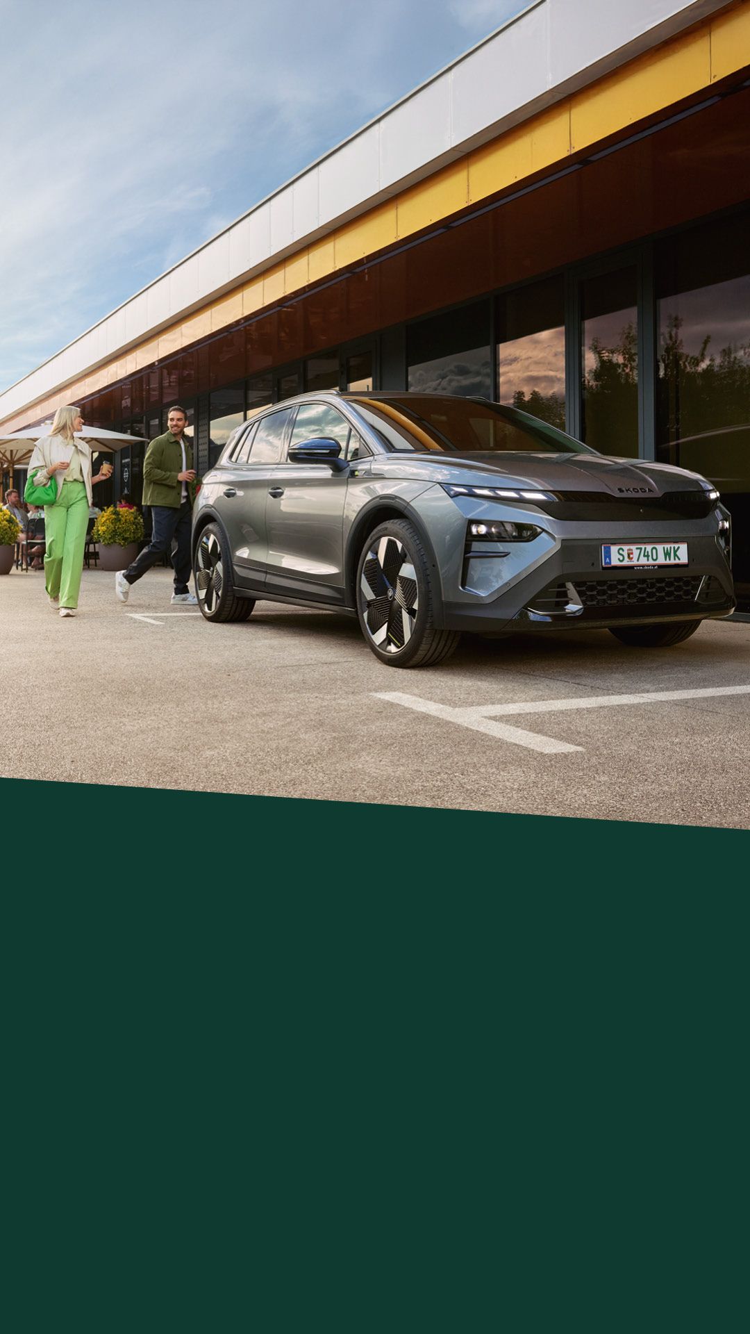 Ein Mann und eine Frau verabschieden sich hinter einem geparkten, grauen Škoda Elroq.