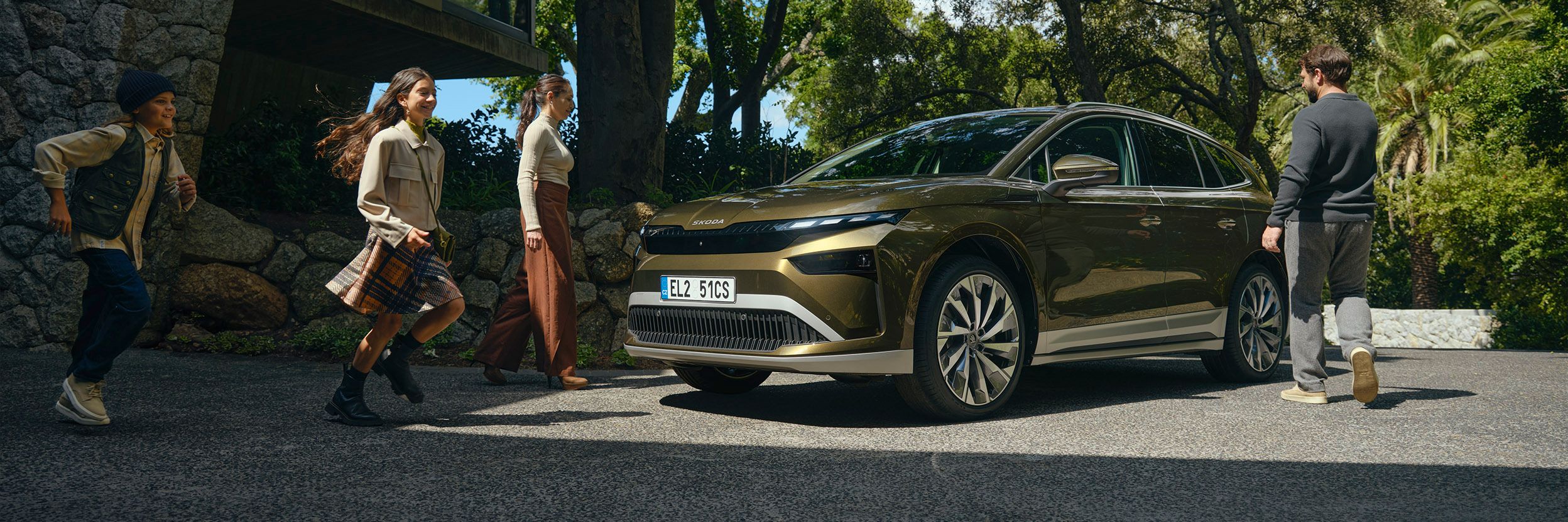 Eine Familie steht auf einem Parkplatz neben einem Haus und sie gehen auf einen grünen Škoda Enyaq zu