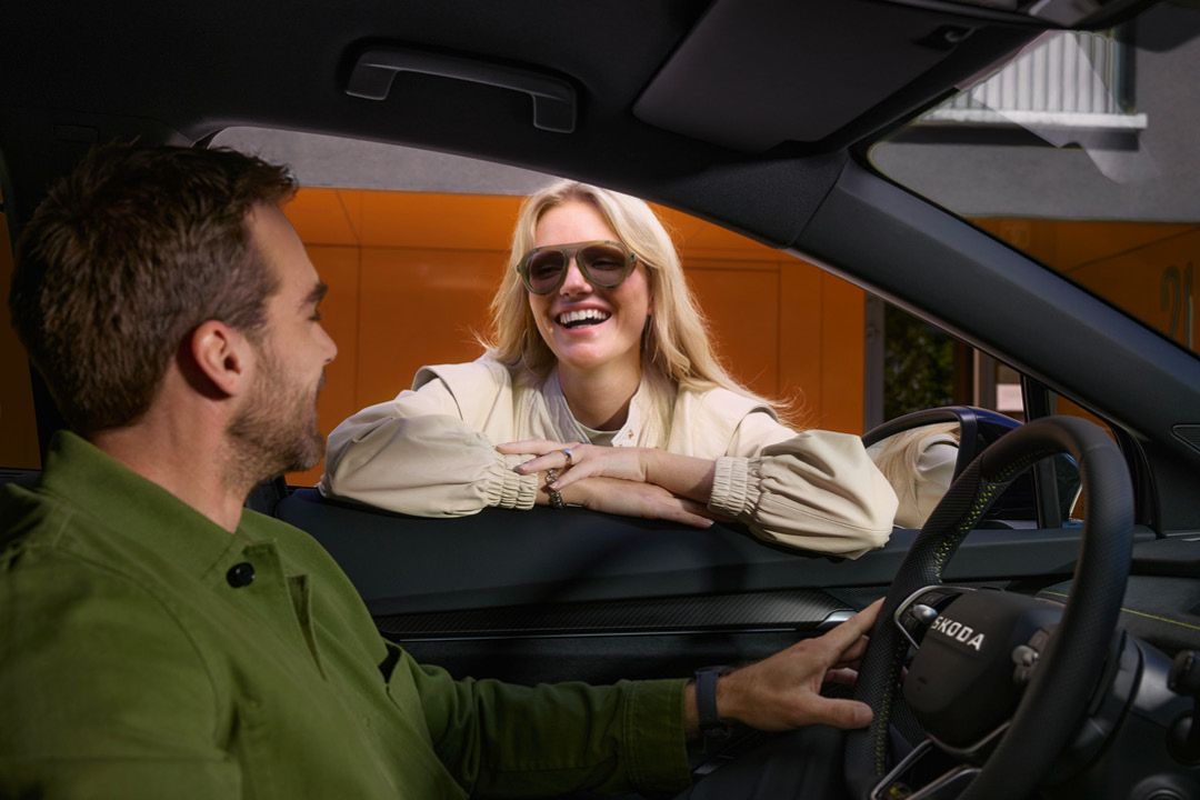 Eine Frau lacht ins offene Fahrerseitenfenster zu einem Mann der in einem Škoda am Steuer sitzt.