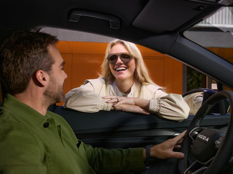 Eine Frau lacht ins offene Fahrerseitenfenster zu einem Mann der in einem Škoda am Steuer sitzt.