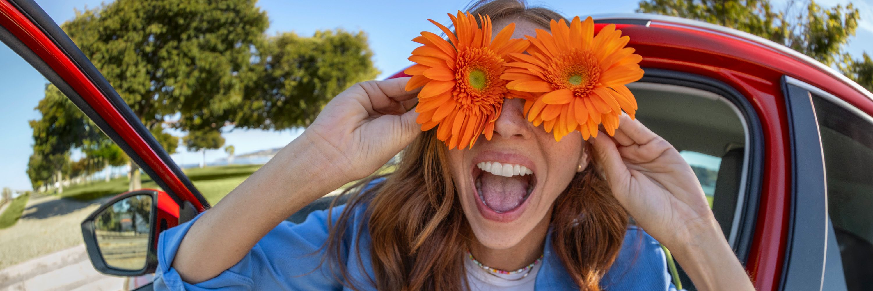 Eine Frau sitzt lachend in einem roten Škoda und hält sich zwei orangefarbene Blumen an die Augen.