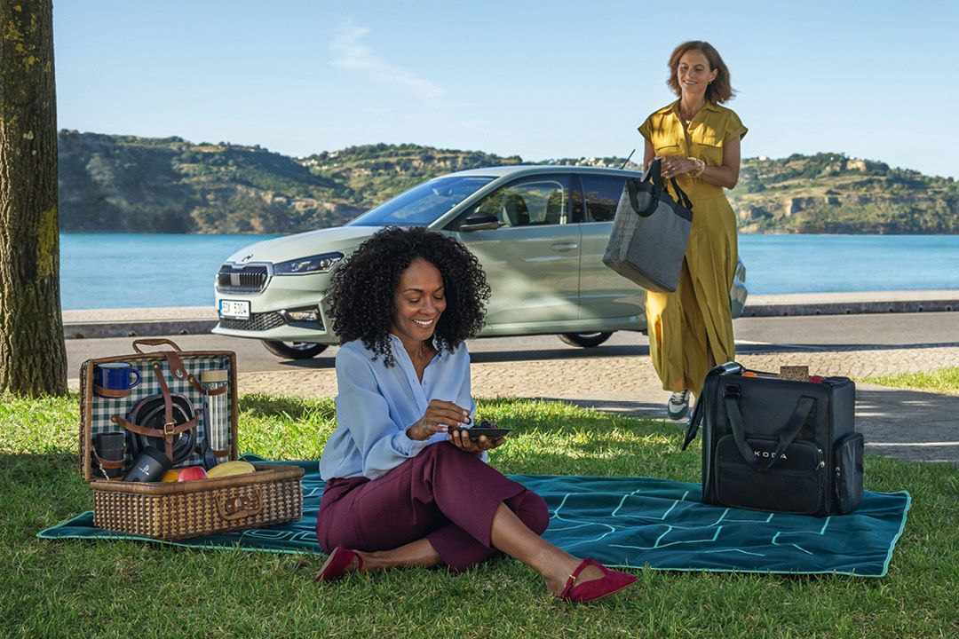 Zwei Frauen sind bei einem Picknick am See. Eine Frau sitzt auf einer Decke und isst, während die andere Frau im gelben Kleid einige Taschen trägt und sich der Decke nähert. Im Hintergrund ist ein geparkter Škoda und eine schöne Seelandschaft sichtbar.