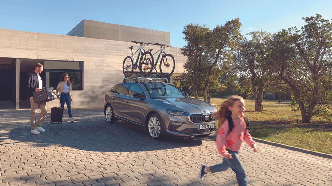 Ein grauer Škoda Scala mit Fahrraddachträger parkt vor einem Haus, die Eltern bereiten sich mit ihrer Tochter auf die Fahrt vor.