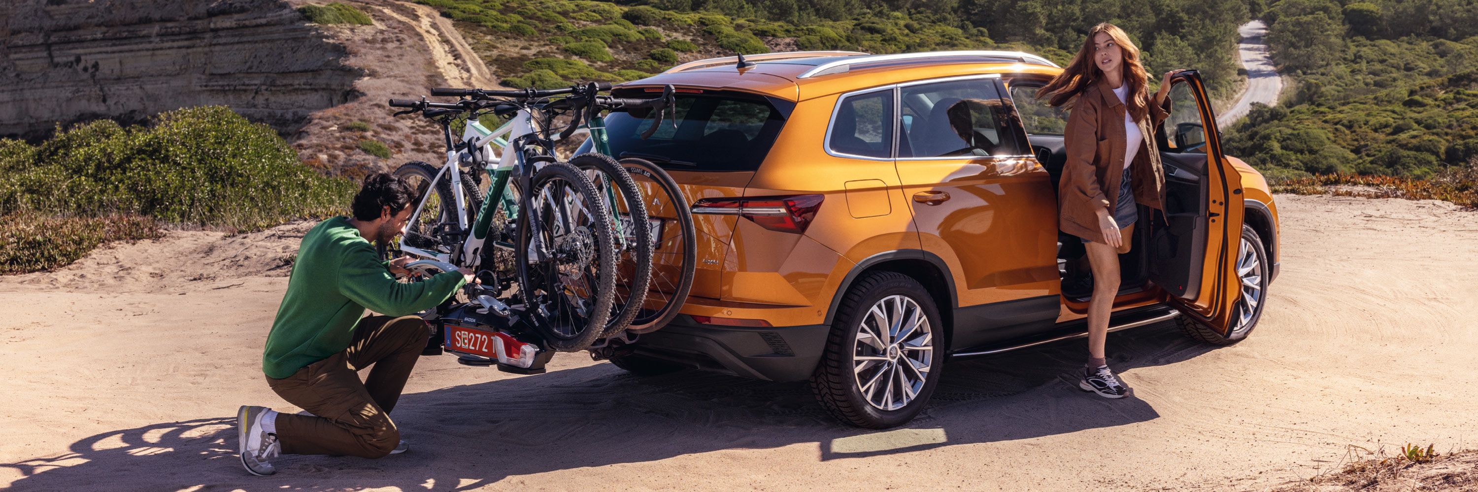 Ein orangefarbener Škoda Kodiaq steht auf einem sandigen Weg. Zwei Fahrräder sind auf einem Fahrradträger am Heck des Autos befestigt. Eine Person in grüner Kleidung überprüft den Fahrradträger, während eine Person in brauner Kleidung aus dem Fahrzeug steigt