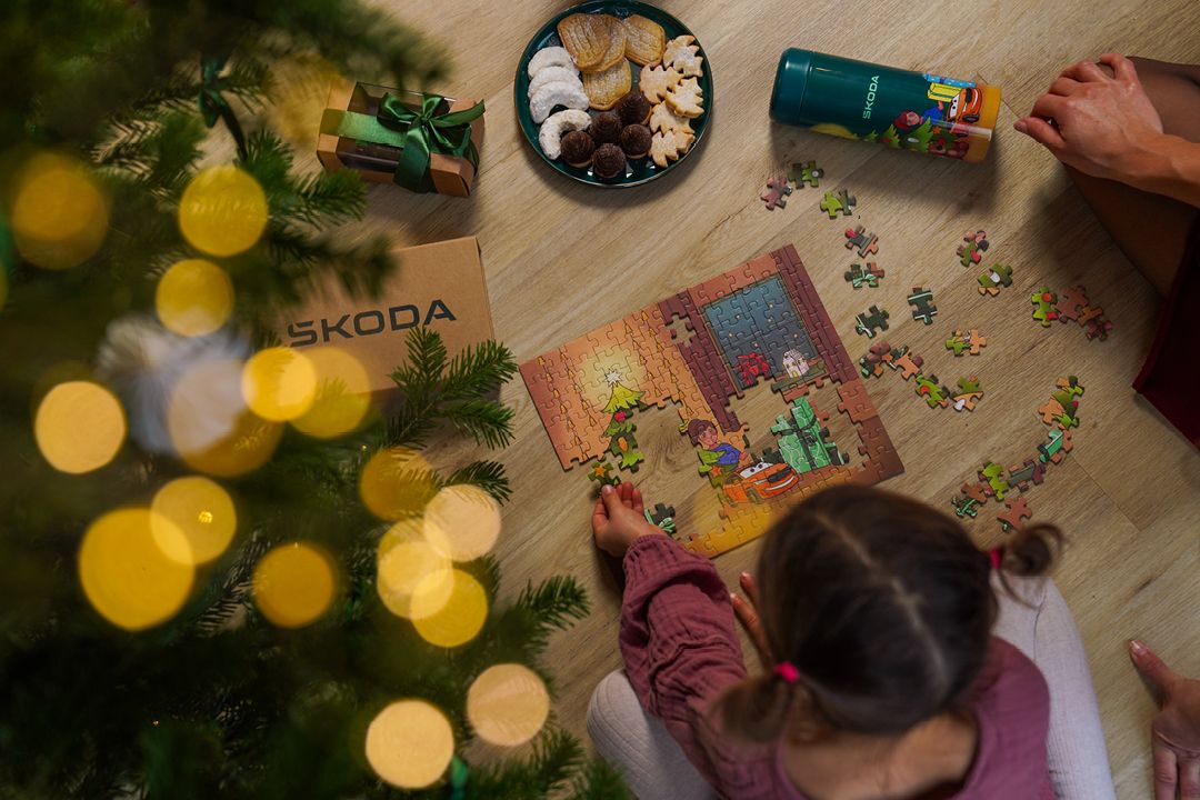 Ein Kind sitzt auf dem Boden und legt ein buntes Škoda Puzzle mit einer weihnachtlichen Szene zusammen. Daneben stehen ein Teller mit Weihnachtsplätzchen, ein Getränkebecher und ein verpacktes Geschenk, während unten links unscharf ein geschmückter Weihnachtsbaum mit Lichtern zu sehen ist. 