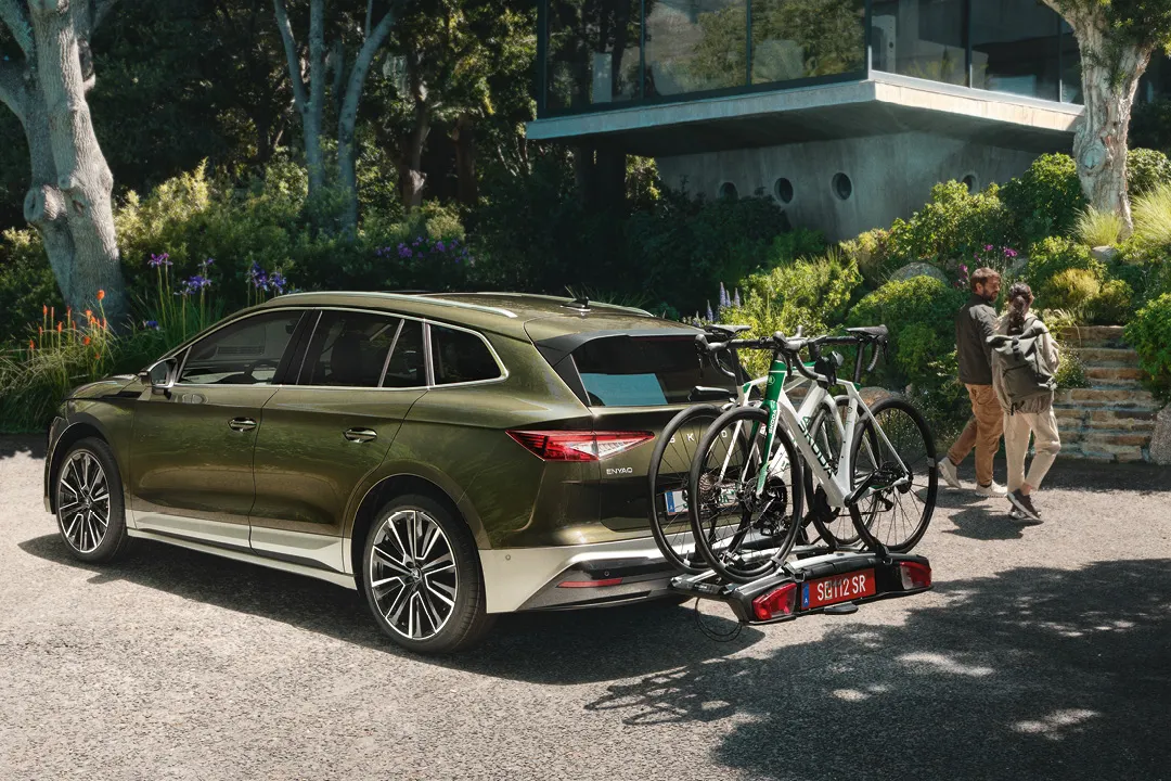Ein grüner Škoda Enyaq mit zwei Fahrrädern auf einem Fahrradträger ist vor einem modernen Haus geparkt. Zwei Personen laufen darauf zu. Die Umgebung ist von Bäumen und Pflanzen umgeben.
